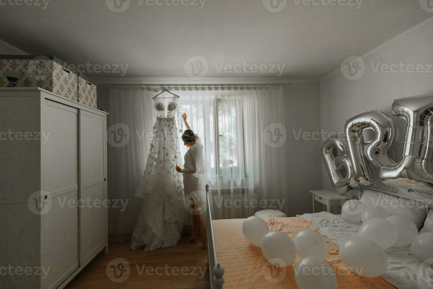 un' bellissimo giovane sposa nel un' bianca sottoveste è in piedi di il finestra, toccante sua nozze vestire. finale preparazioni per il nozze. il sposa è in attesa per il sposo. mattina, sposa. foto