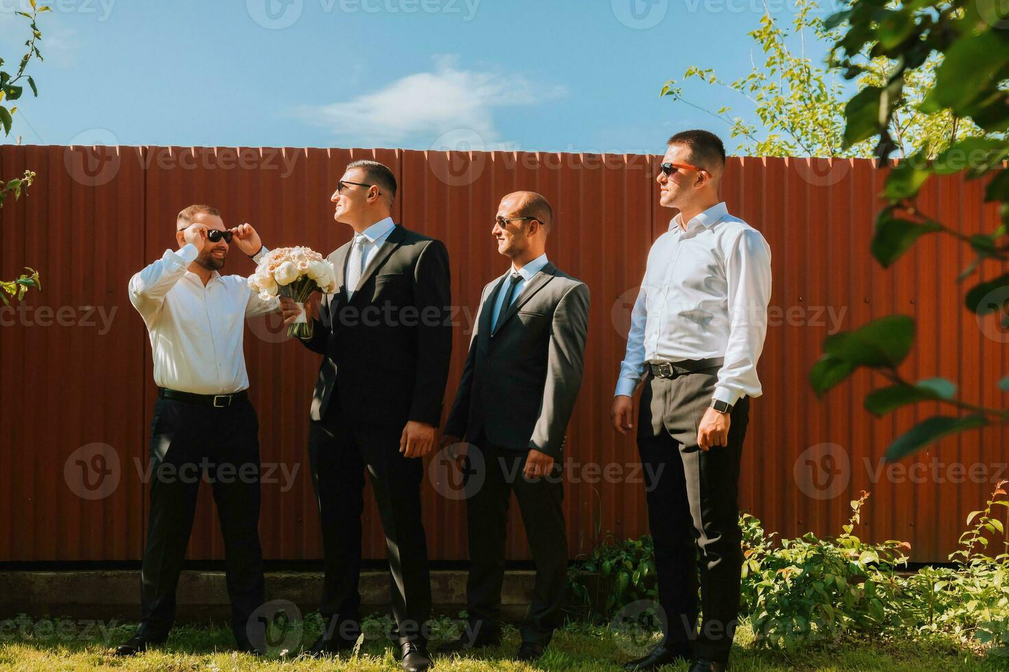 il sposo nel un' nero completo da uomo e bicchieri e il suo elegante amici indossare bianca camicie e nero pantaloni e bicchieri siamo in piedi nel il Giardino dietro la casa nel il giardino. il sposo è Tenere un' mazzo. foto