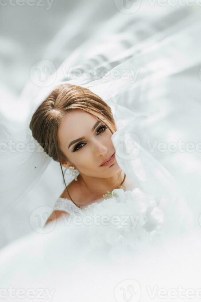 un' sposa nel un' nozze vestito con un' lungo treno e un' velo detiene un' nozze mazzo di Rose mentre seduta sotto un' velo. foto a partire dal sopra