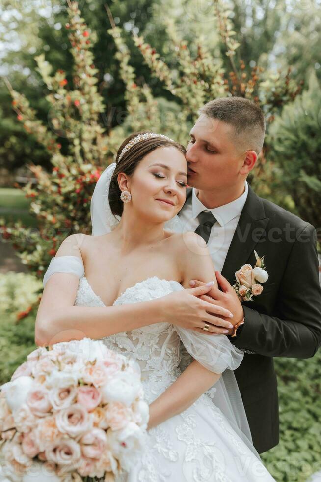 sposo e sposa baci nel il parco. un' coppia di Novelli sposi, il sposa e sposo, a un' nozze nel natura, un' verde foresta, baci, foto ritratto. nozze coppia