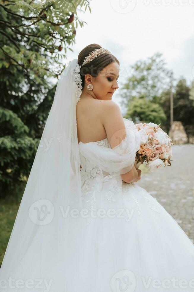 posteriore Visualizza ritratto di bellissimo sposa nel bianca nozze vestito con lungo treno con moderno acconciatura e velo a piedi nel giardino. nozze concetto foto