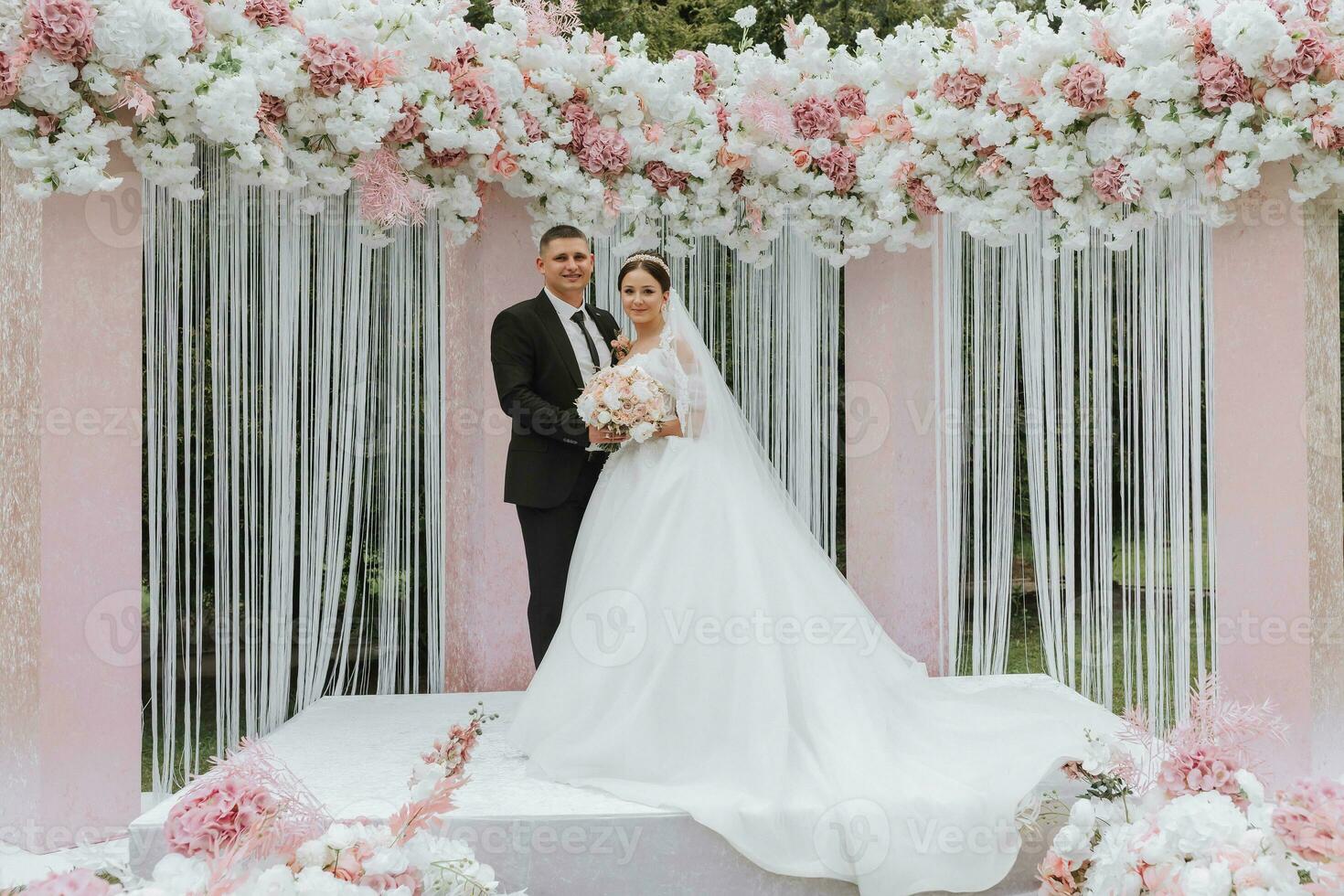 attraente sposa e sposo a il cerimonia su loro nozze giorno con un arco fatto di rosa e bianca fiori. bellissimo Novelli sposi, un' giovane donna nel un' bianca vestito con un' lungo treno, uomini nel un' nero completo da uomo. foto