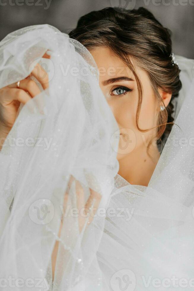 un' sbalorditivo giovane sposa pose di il finestra Tenere sua velo nel sua mani. un' bellissimo donna di caucasico aspetto nel un elegante bianca accappatoio e lussuoso acconciatura. nozze trucco foto