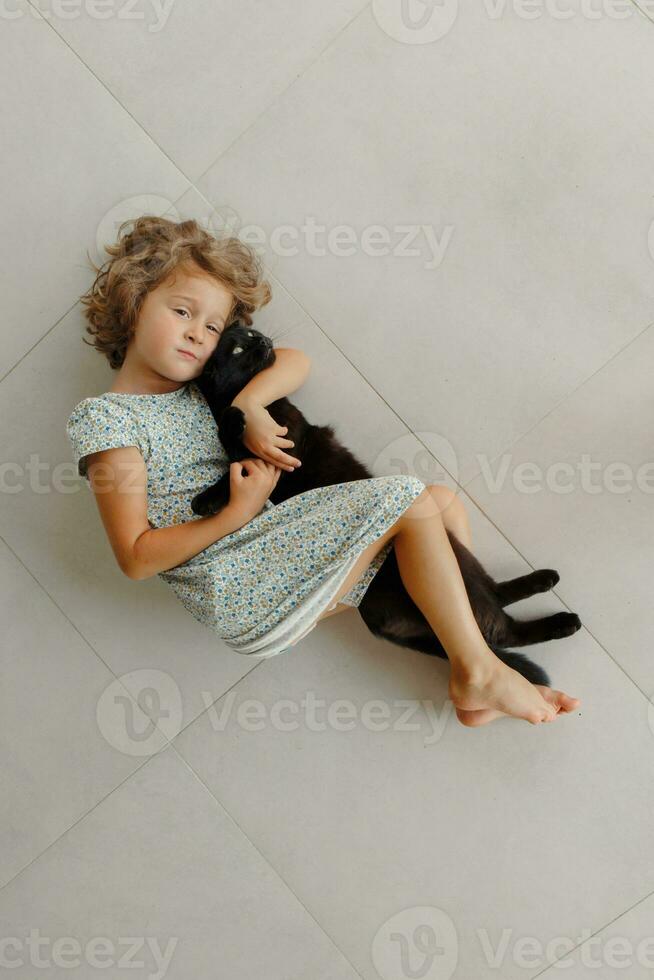un' contento bambino è dire bugie su il veranda con il suo preferito nero gatto. un' poco ragazza riposa su il pavimento di il veranda su un' soleggiato estate giorno. un' contento e spensierato infanzia. foto