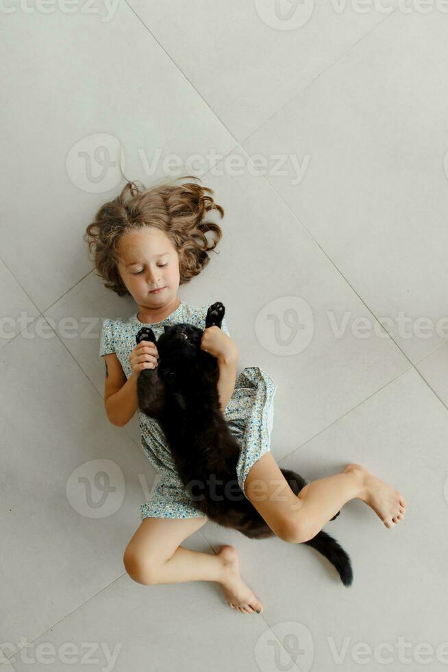 un' contento bambino è dire bugie su il veranda con il suo preferito nero gatto. un' poco ragazza riposa su il pavimento di il veranda su un' soleggiato estate giorno. un' contento e spensierato infanzia. foto