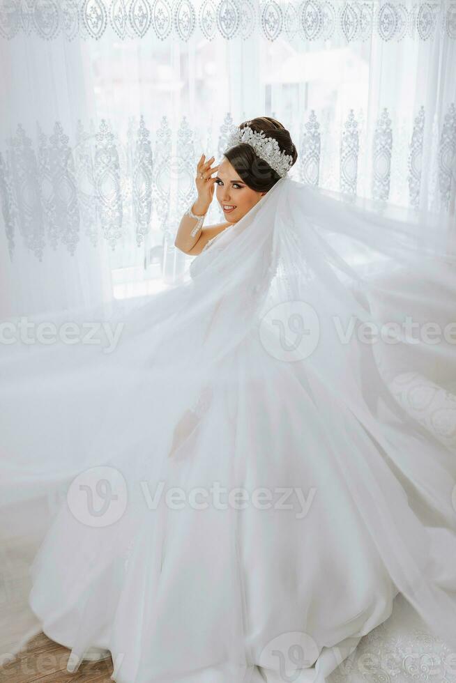 ritratto di un' sposa con un' bellissimo acconciatura e tiara su sua testa e naturale trucco in posa nel un' bianca accappatoio nel sua camera. il bellezza di il ragazza. un' lungo bianca velo foto