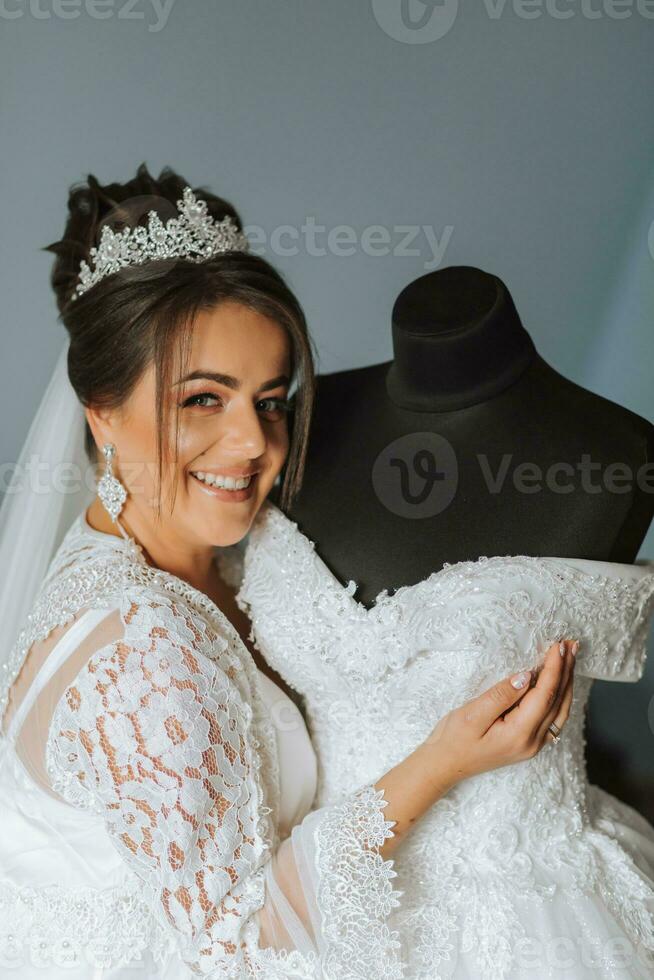 un' contento sposa è preparazione per sua lussuoso nozze a casa, Tenere sua bellissimo vestito nel sua mani. ritratto di un' donna con professionale capelli e trucco e un' Sorridi nel un' accappatoio foto