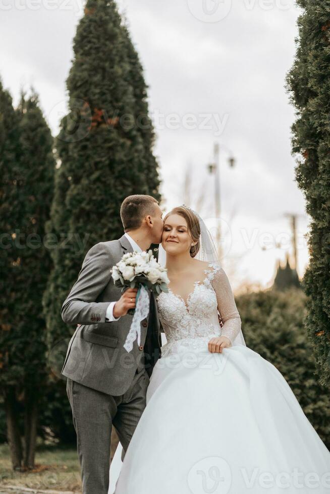 elegante sposa e sposo teneramente abbraccio e bacio. bellissimo sposa con moderno mazzo Abbracciare una persona elegante sposo nel autunno parco. sensuale romantico momento. nozze. elegante e Bellissima. foto