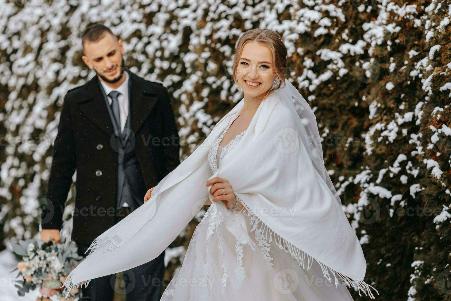 ritratto di contento Novelli sposi su il sfondo di innevato alberi. il sposo abbracci il sposa nel il inverno parco. sorridente sposa nel nozze vestito e bianca poncho. il sposo è vestito nel un' nero cappotto. foto