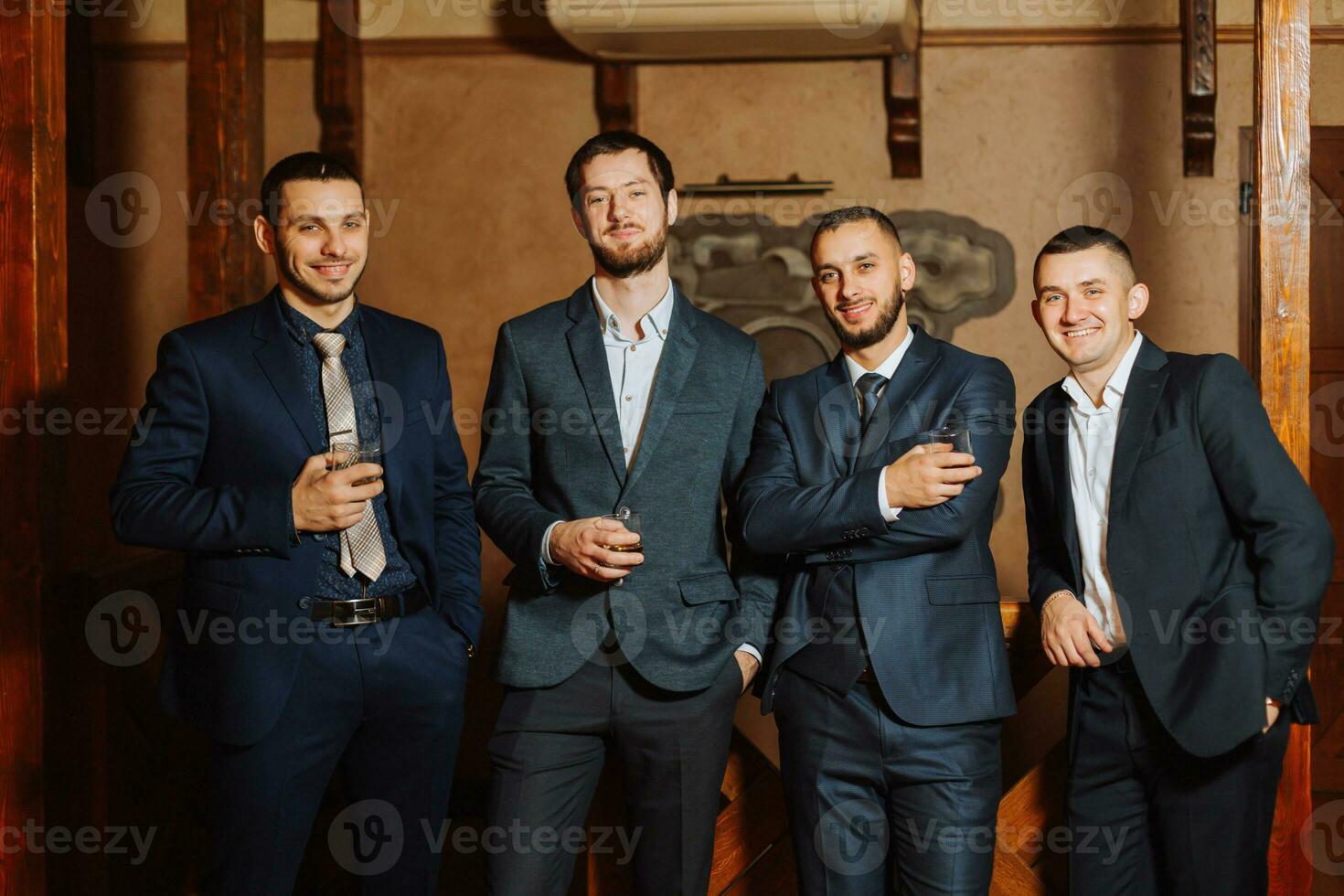 il sposo e il suo amici nel elegante tute bevanda whisky nel il Hotel camera, il mattina prima il nozze preparativi. ritratto di uomini potabile whisky foto