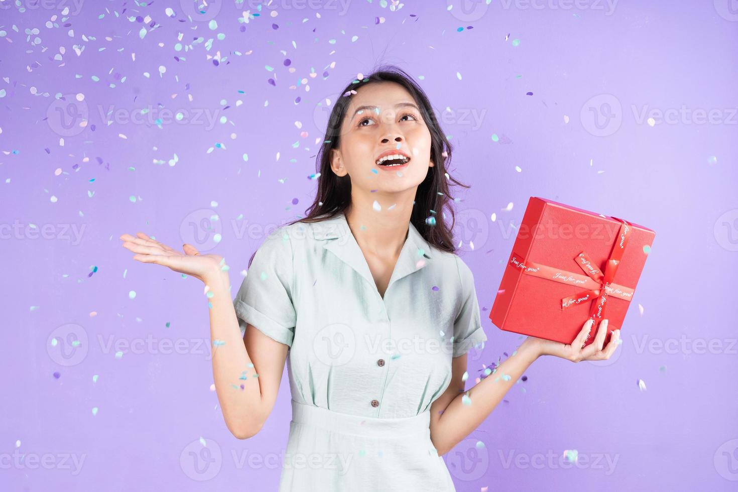 ritratto di bella ragazza in mano con scatola regalo rossa e festeggia sotto la pioggia di coriandoli, isolato su sfondo viola foto