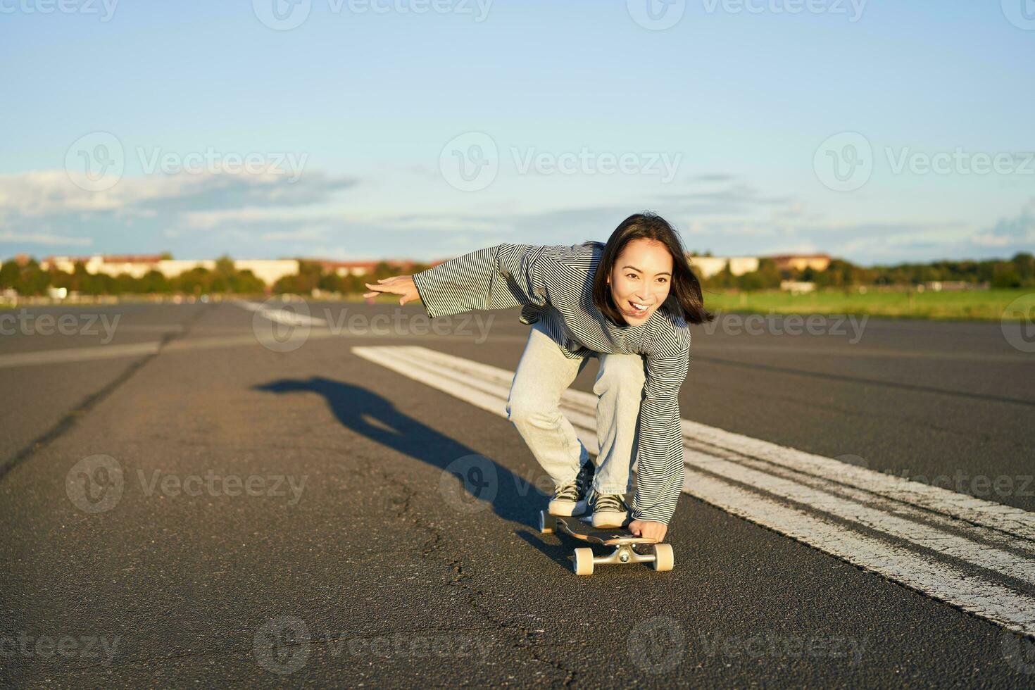 spensierato pattinatore ragazza su sua skateboard, equitazione longboard su un vuoto strada, Tenere mani lateralmente e ridendo foto