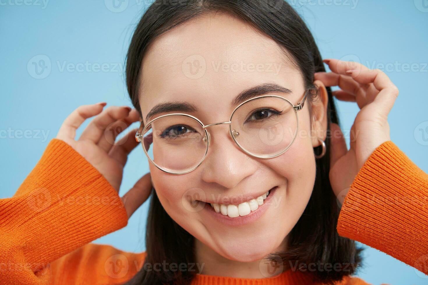 vicino su ritratto di bellissimo giovane asiatico donna nel occhiali, sorridente e guardare felice, provando nuovo occhiali a ottici negozio, sta al di sopra di blu sfondo foto