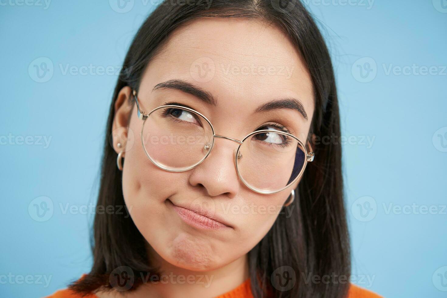 ritratto di giapponese donna nel occhiali, sembra premuroso, medita, pensiero con grave viso, in piedi al di sopra di blu sfondo foto