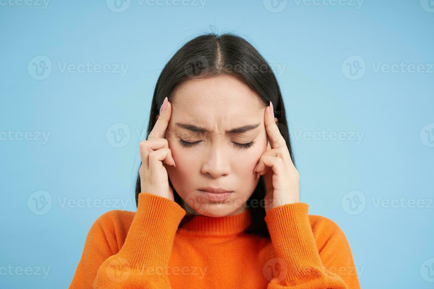 vicino su ritratto di asiatico donna con emicrania, tocchi sua testa e massaggio templi, ha male alla testa, sta al di sopra di blu sfondo foto