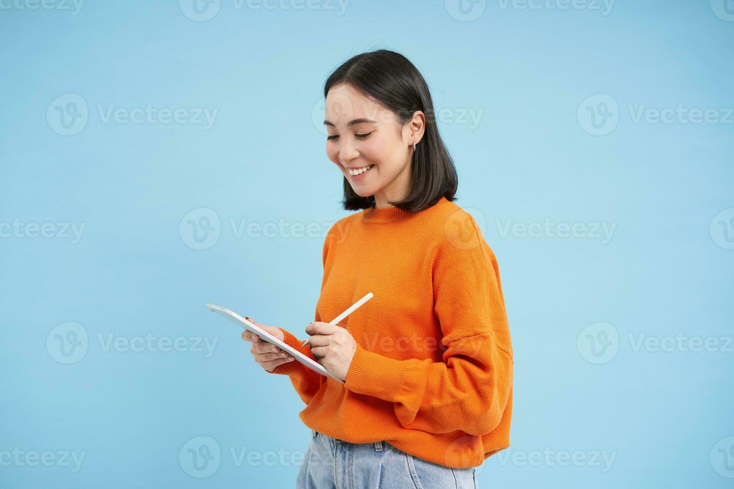 bellissimo moderno Cinese donna con digitale tavoletta e matita, guardare a schermo, scrittura giù Appunti, fare compiti a casa, disegno su gadget, blu sfondo foto