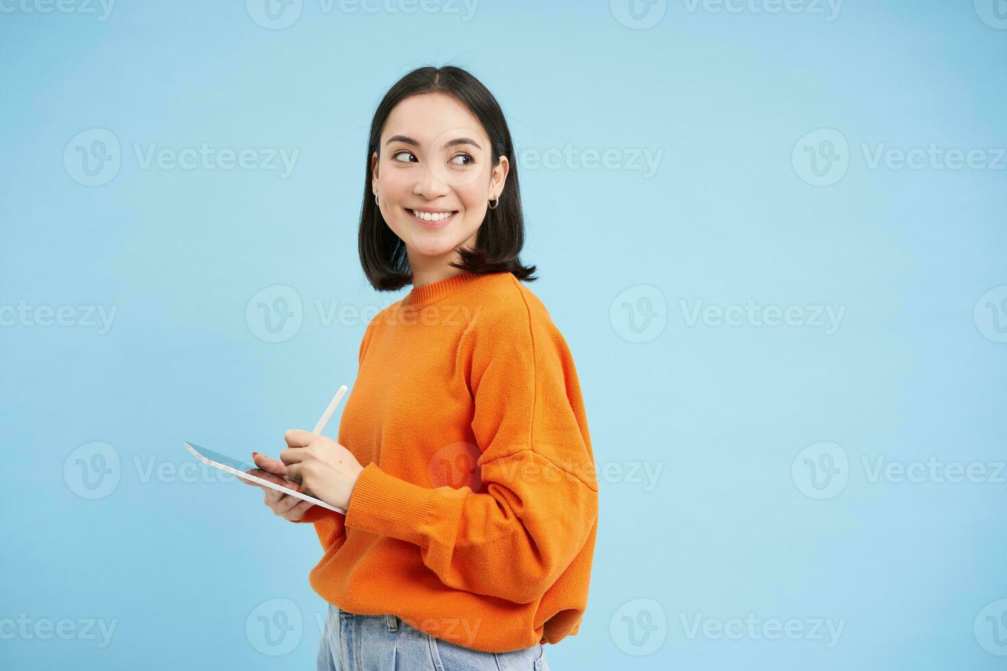 moderno bellissimo asiatico donna con digitale tavoletta e matita, assunzione Appunti, scrittura su sua gadget, fare compiti a casa, Lavorando, in piedi al di sopra di blu sfondo foto