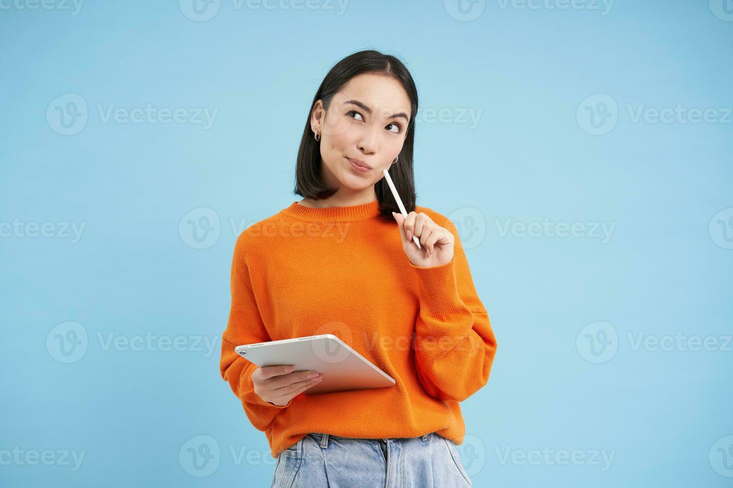 moderno bellissimo asiatico donna con digitale tavoletta e matita, assunzione Appunti, scrittura su sua gadget, fare compiti a casa, Lavorando, in piedi al di sopra di blu sfondo foto