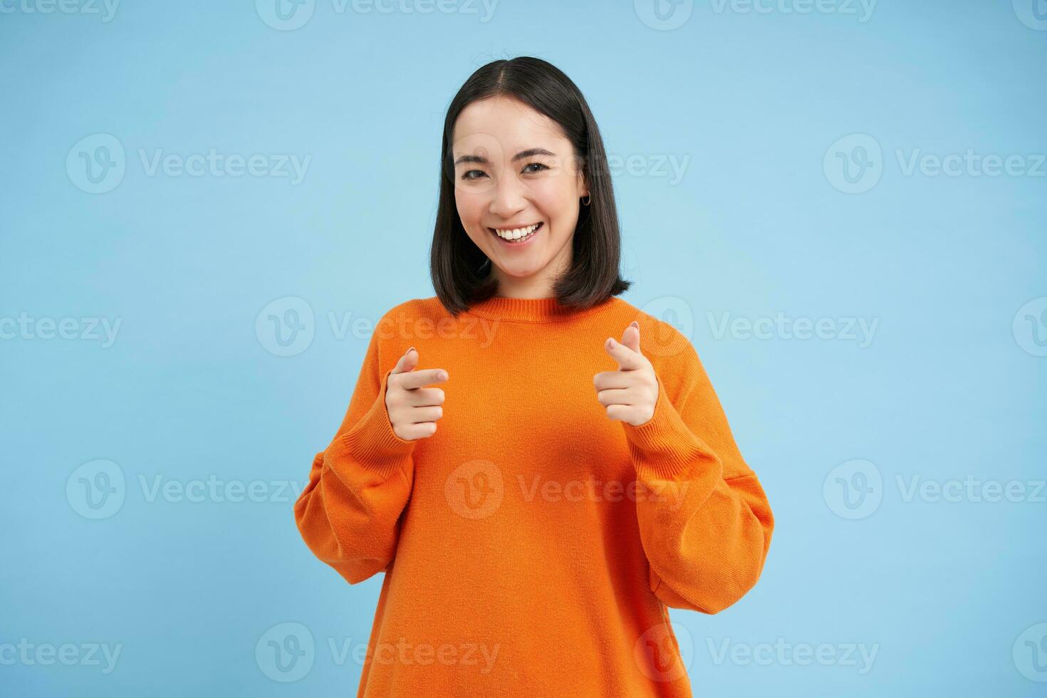 voi avuto questo, bene lavoro. entusiasta coreano donna, punti a telecamera con dito pistole e sorrisi, incoraggia, lodi, sta al di sopra di blu sfondo foto