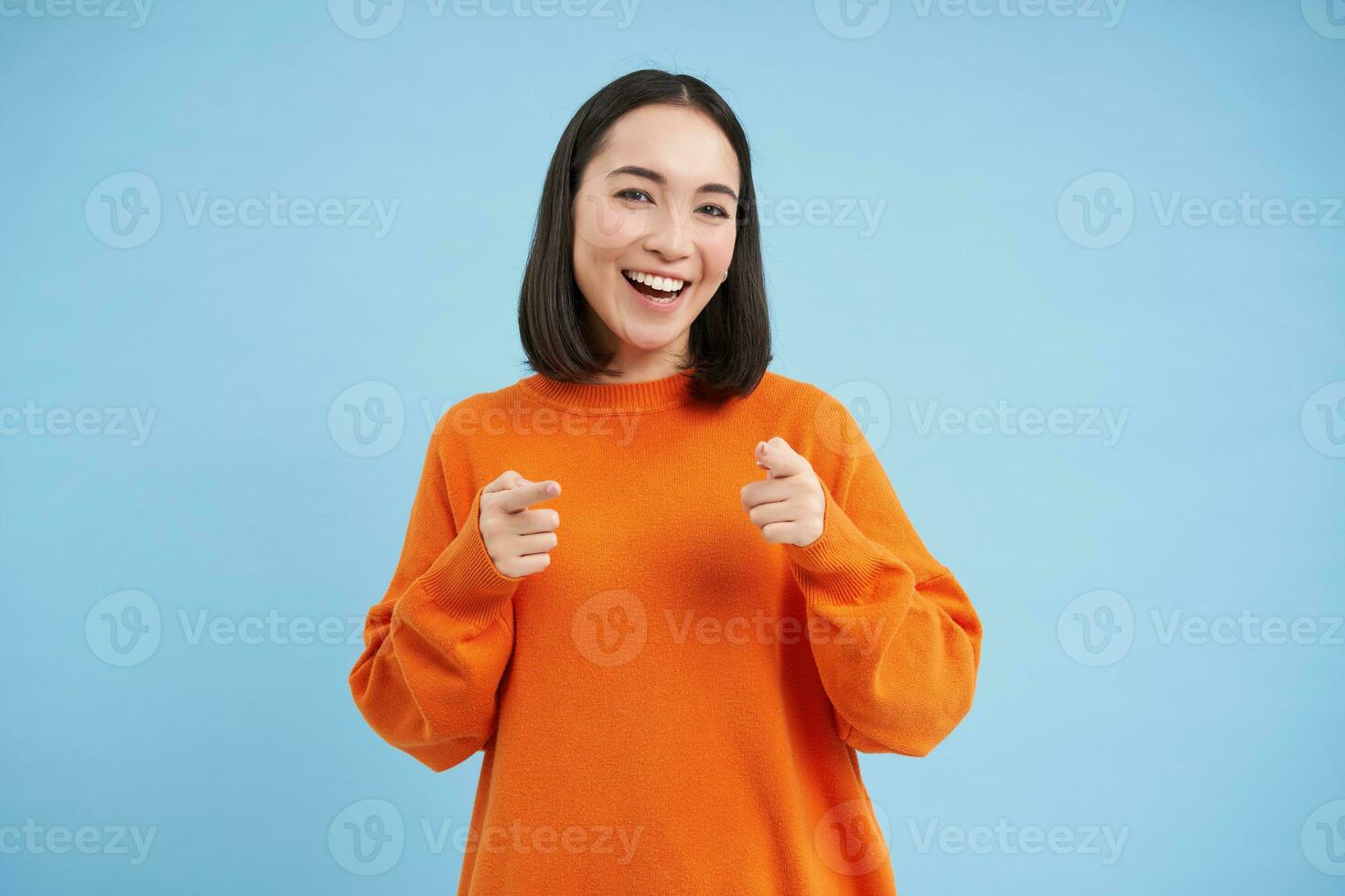 voi avuto questo, bene lavoro. entusiasta coreano donna, punti a telecamera con dito pistole e sorrisi, incoraggia, lodi, sta al di sopra di blu sfondo foto