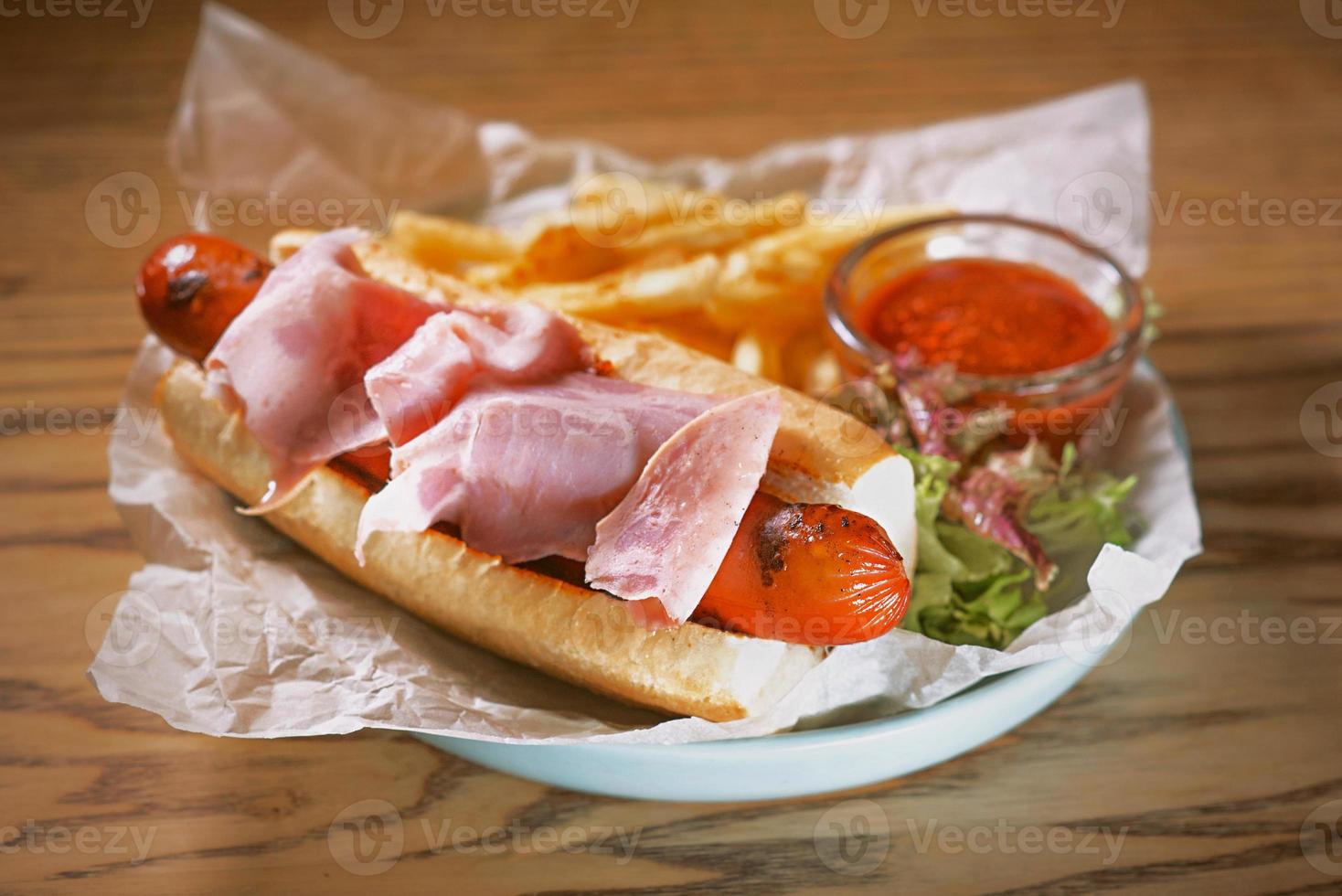 piatto con hot dog con salsiccia e prosciutto, salsa di pomodoro ketchup e patatine fritte sul tavolo foto