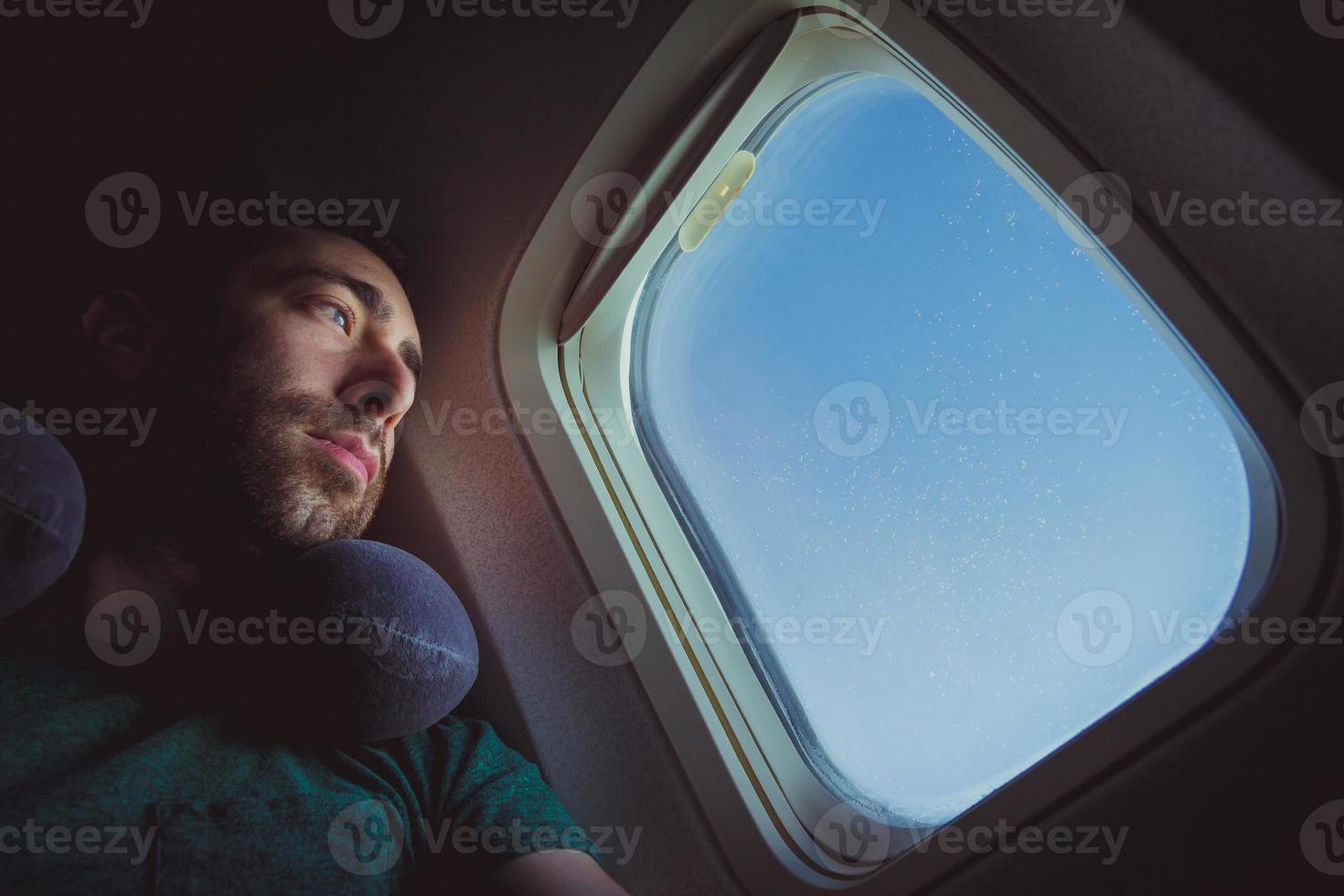 uomo pensieroso con cuscino per il collo che guarda fuori attraverso il finestrino di un aeroplano foto