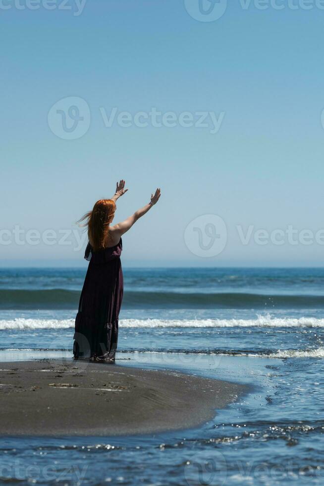 donna nel lungo vestito in piedi su sabbioso spiaggia con sua braccia sollevato alto, raggiungendo su per cielo e mare foto