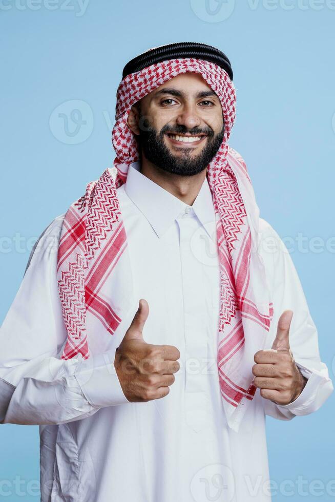 allegro musulmano uomo indossare bianca thobe e copricapo mostrando pollici su studio ritratto. sorridente arabo persona in posa con positivo gesto, in mostra arabo cultura e guardare a telecamera foto