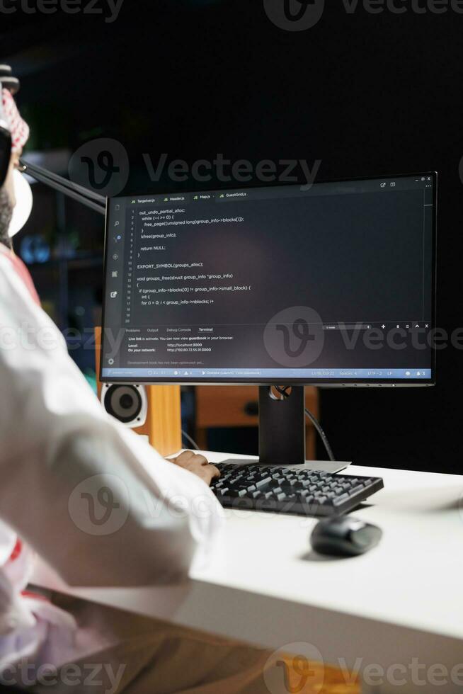 musulmano tipo si siede a scrivania e codici su computer, dimostrando il App sviluppo processi. sopra la spalla tiro di pc tenere sotto controllo esibendo in esecuzione algoritmi prodotta di arabo uomo vestito tradizionalmente. foto