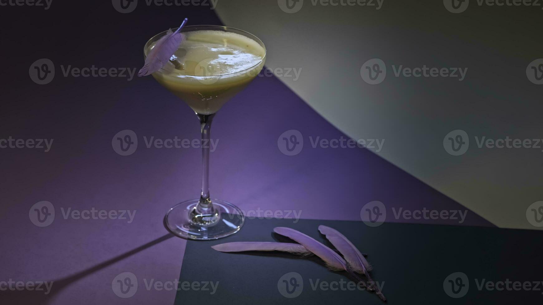 rinfrescante alcolizzato bevanda nel Martini bicchiere. azione filmato. vicino su di bevanda servito su lilla e bianca superficie e decorato con piccolo viola piume. foto