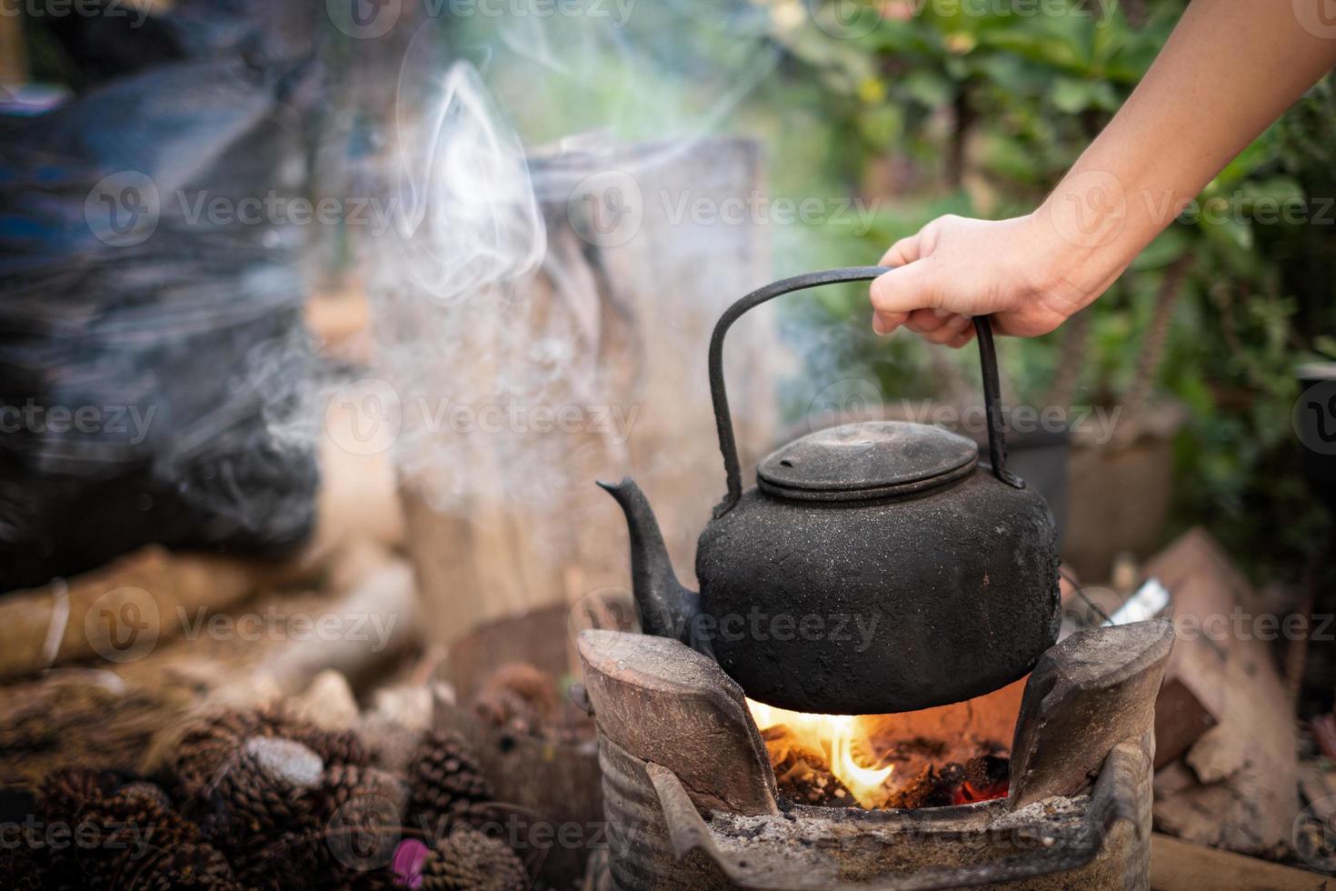 vicino la mano che tiene l'acqua bollire il vecchio bollitore sul fuoco con una stufa a carbone a sfondo sfocato foto