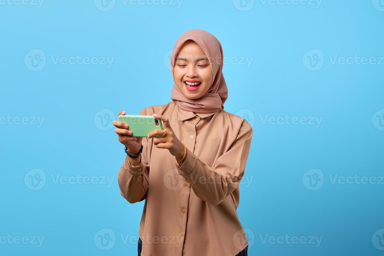 ritratto di allegra giovane donna asiatica che gioca al videogioco sul telefono cellulare su sfondo blu foto