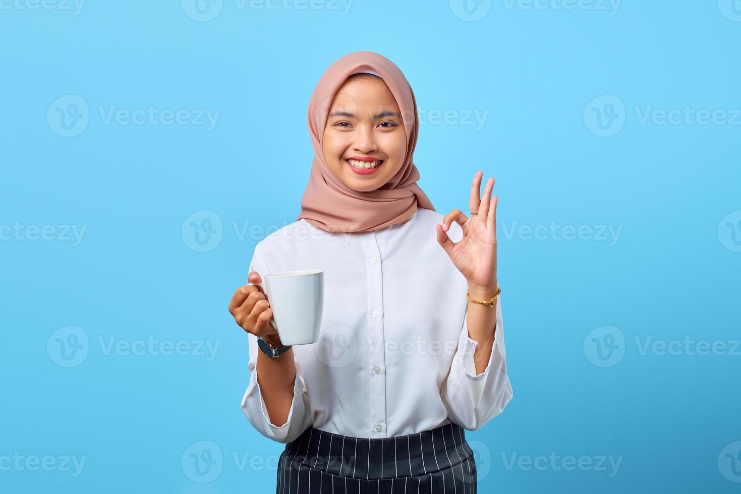 ritratto di giovane donna asiatica sorridente che mostra il segno giusto e che tiene tazza su sfondo blu foto