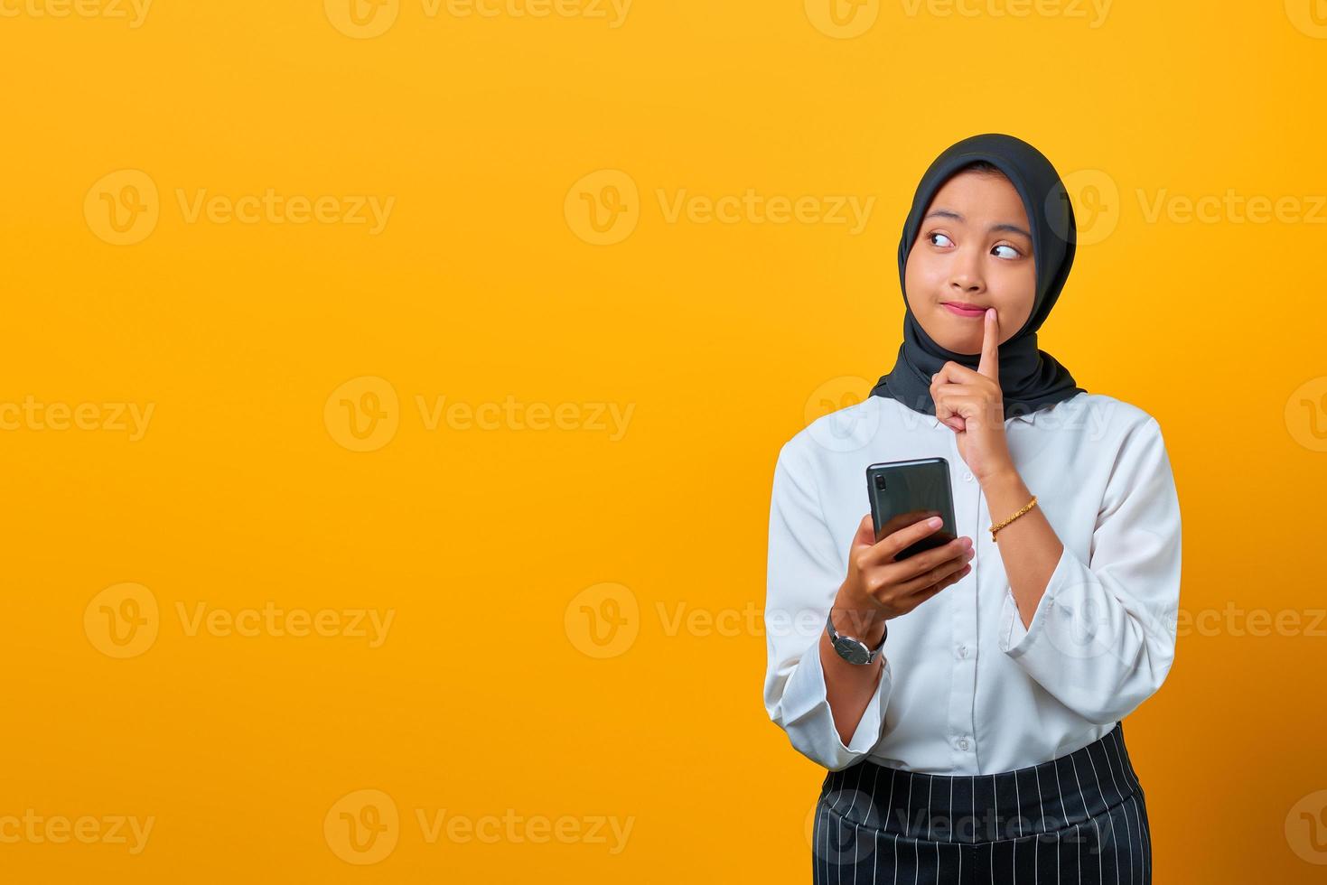 ritratto di giovane donna asiatica pensierosa che tiene il telefono cellulare e la mano sulle labbra su sfondo giallo foto