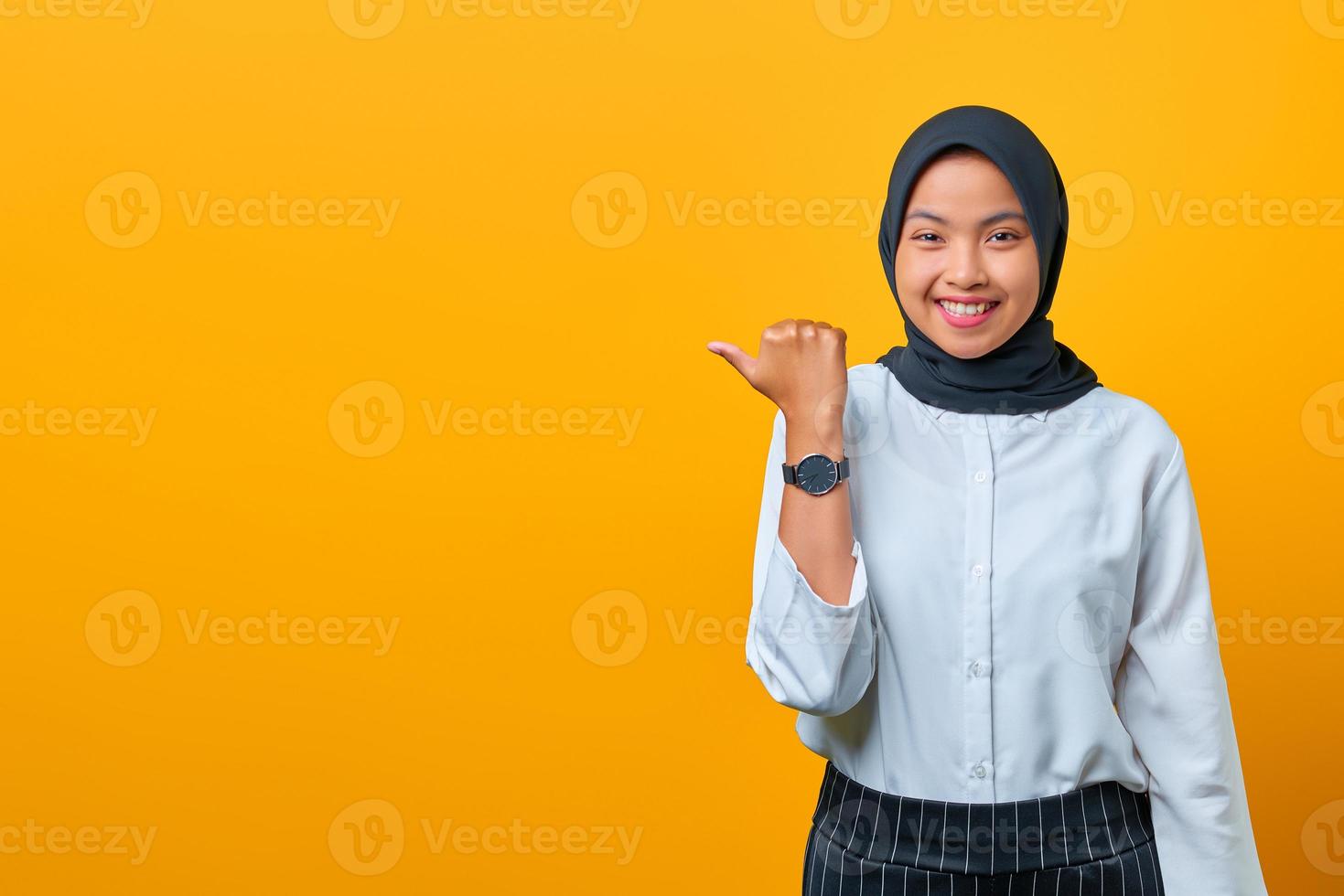 sorridente giovane donna asiatica che punta il dito sullo spazio della copia e guarda la telecamera su sfondo giallo foto