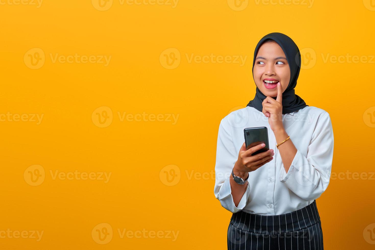 ritratto di giovane donna asiatica attraente che tiene il telefono cellulare e la mano sulle labbra su sfondo giallo foto