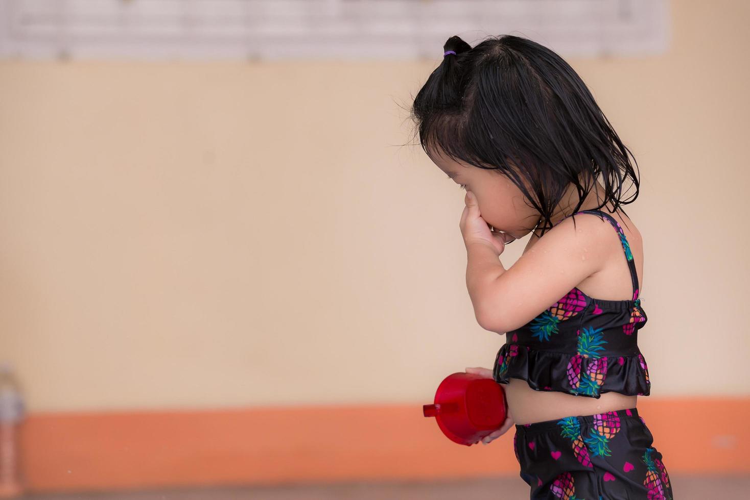 la ragazza carina si è bagnata giocando in acqua davanti alla casa. candido bambino accarezzava le gocce d'acqua dal suo viso, la sua mano destra tenendo la tazza di plastica rossa. il bambino di vista laterale stava portando il costume da bagno nero. foto