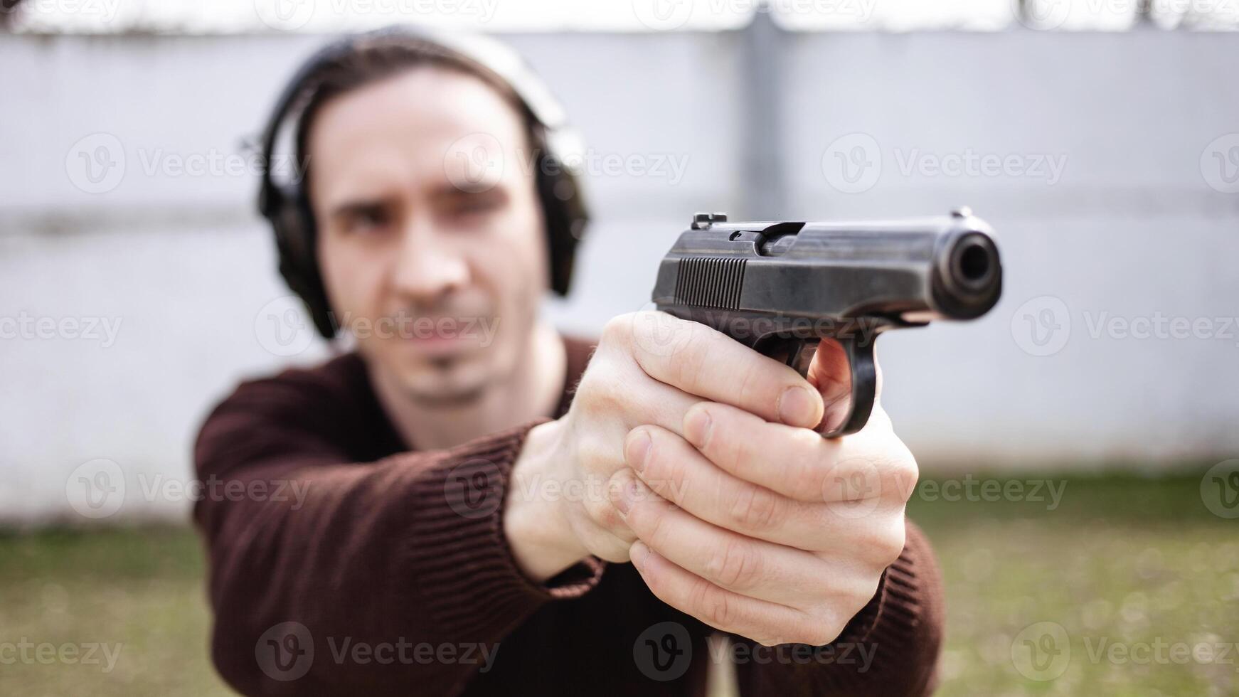 un' uomo è mirando per un' pistola. un' uomo indossare protettivo cuffia. all'aperto pneumatico tiro gamma. passatempo. arma contro il bianca parete foto