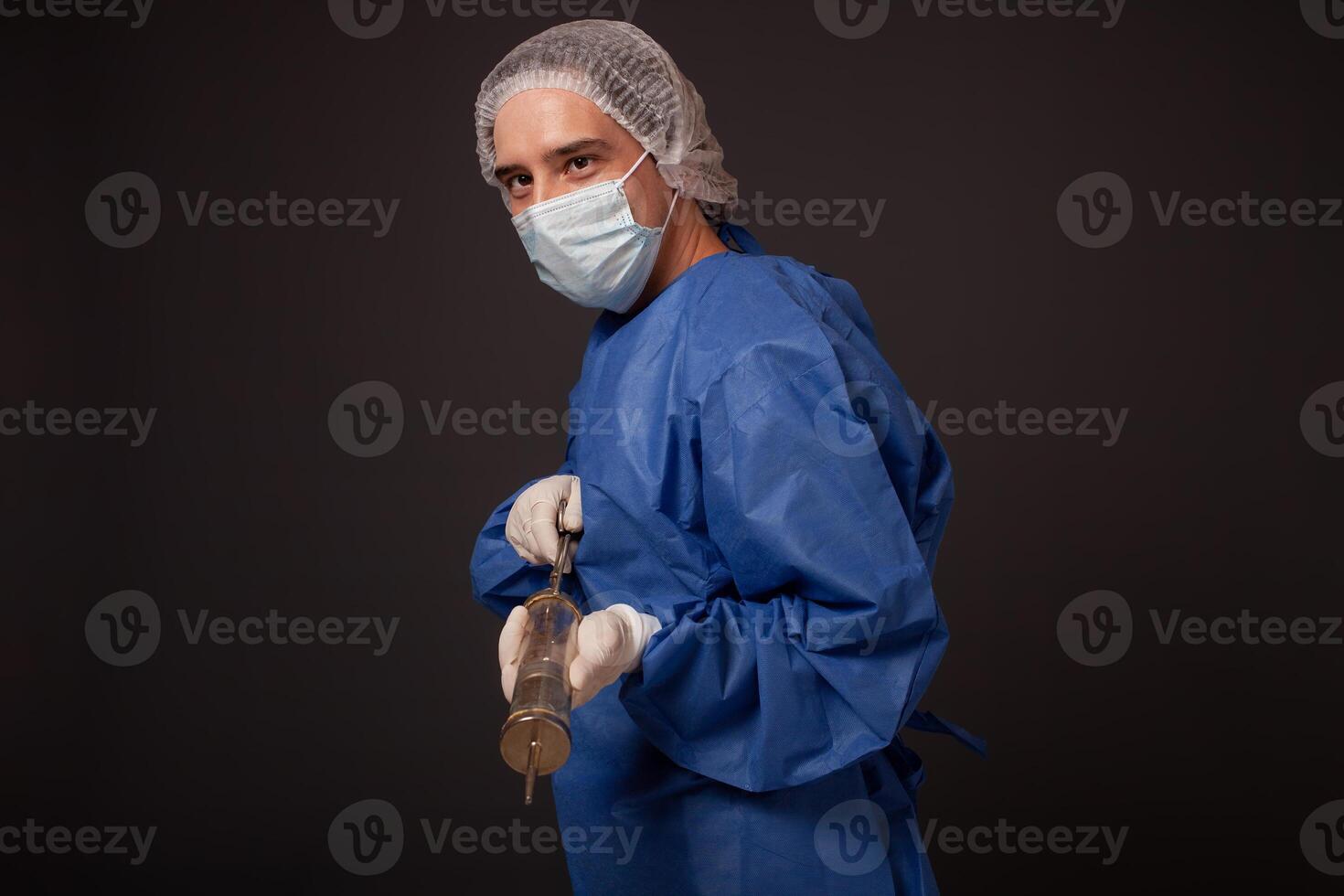 un' maschio medico con un' terribile vecchio siringa nel il suo mani. il medico nel un' medico maschera, cappello, accappatoio e guanti. un' terribile. il medico spaventa con un' enorme siringa con un' di spessore ago. foto