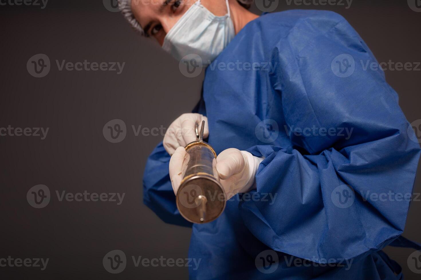 un' maschio medico con un' terribile vecchio siringa nel il suo mani. il medico nel un' medico maschera, cappello, accappatoio e guanti. un' terribile. il medico spaventa con un' enorme siringa con un' di spessore ago. foto