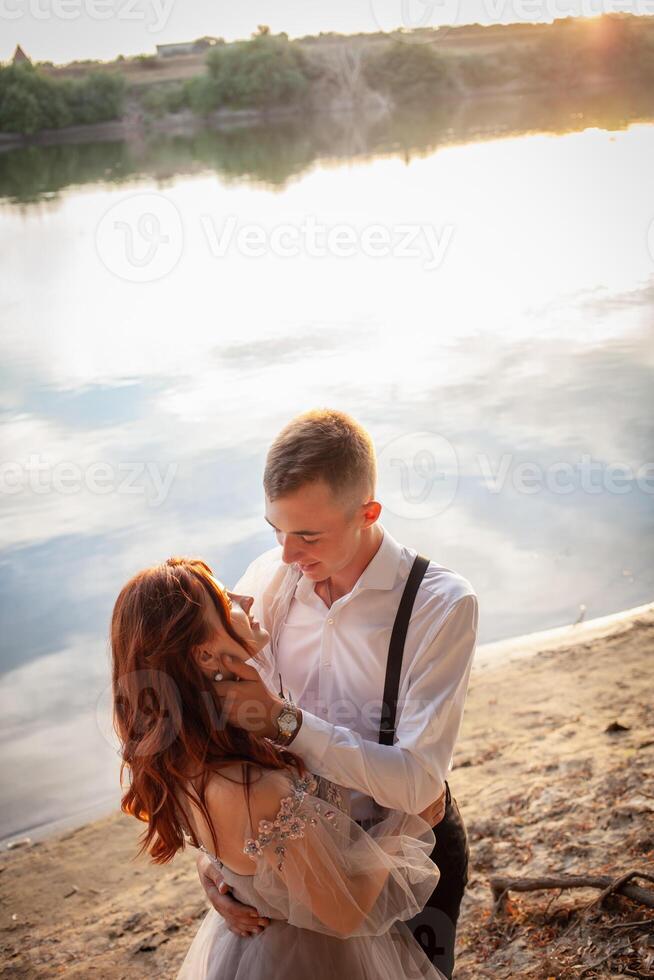 un' bellissimo nozze coppia su il Riva del fiume a tramonto. un' donna nel un' grigio vestito con fiori, un' uomo nel un' completo da uomo e giarrettiere abbraccio ogni Altro, amore. Gli amanti su un' camminare foto