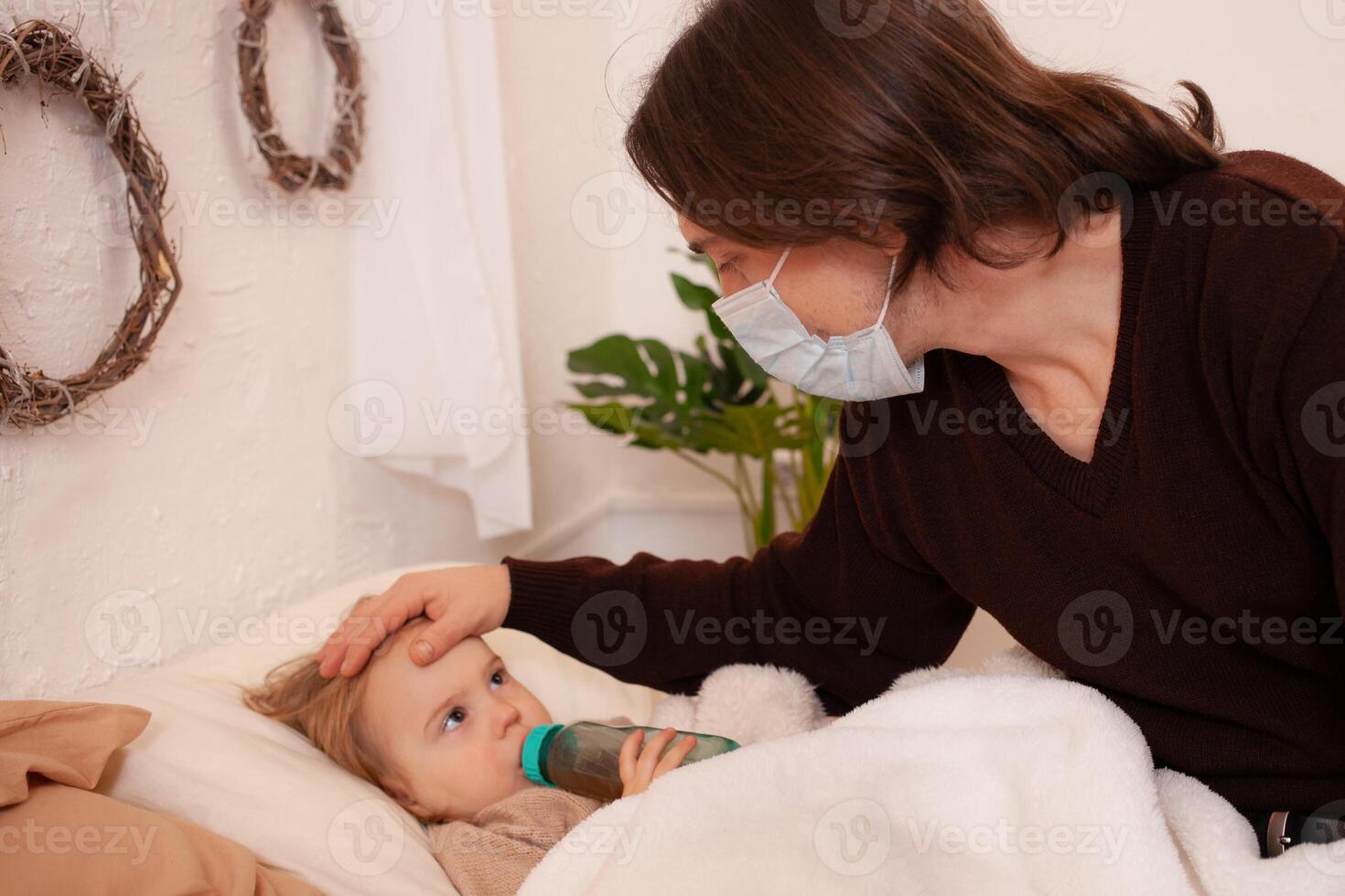 il bambino è malato, il padre nel un' maschera controlli il temperatura di il suo figlia. influenza, freddo, coronavirus, covid, casa quarantena. il bambino è malato e bugie su il letto, il uomo è assunzione cura foto