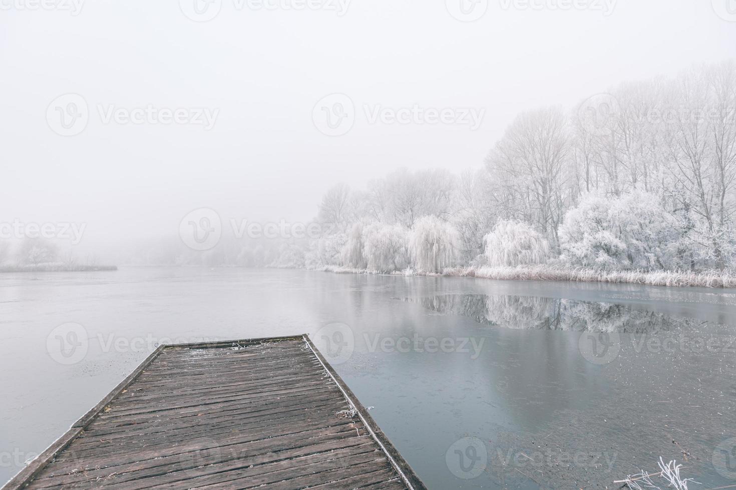 paesaggio invernale serale. molo in legno su un bellissimo lago ghiacciato. alberi con gelo, calmo scenario invernale stagionale. vista pacifica e bianca foto