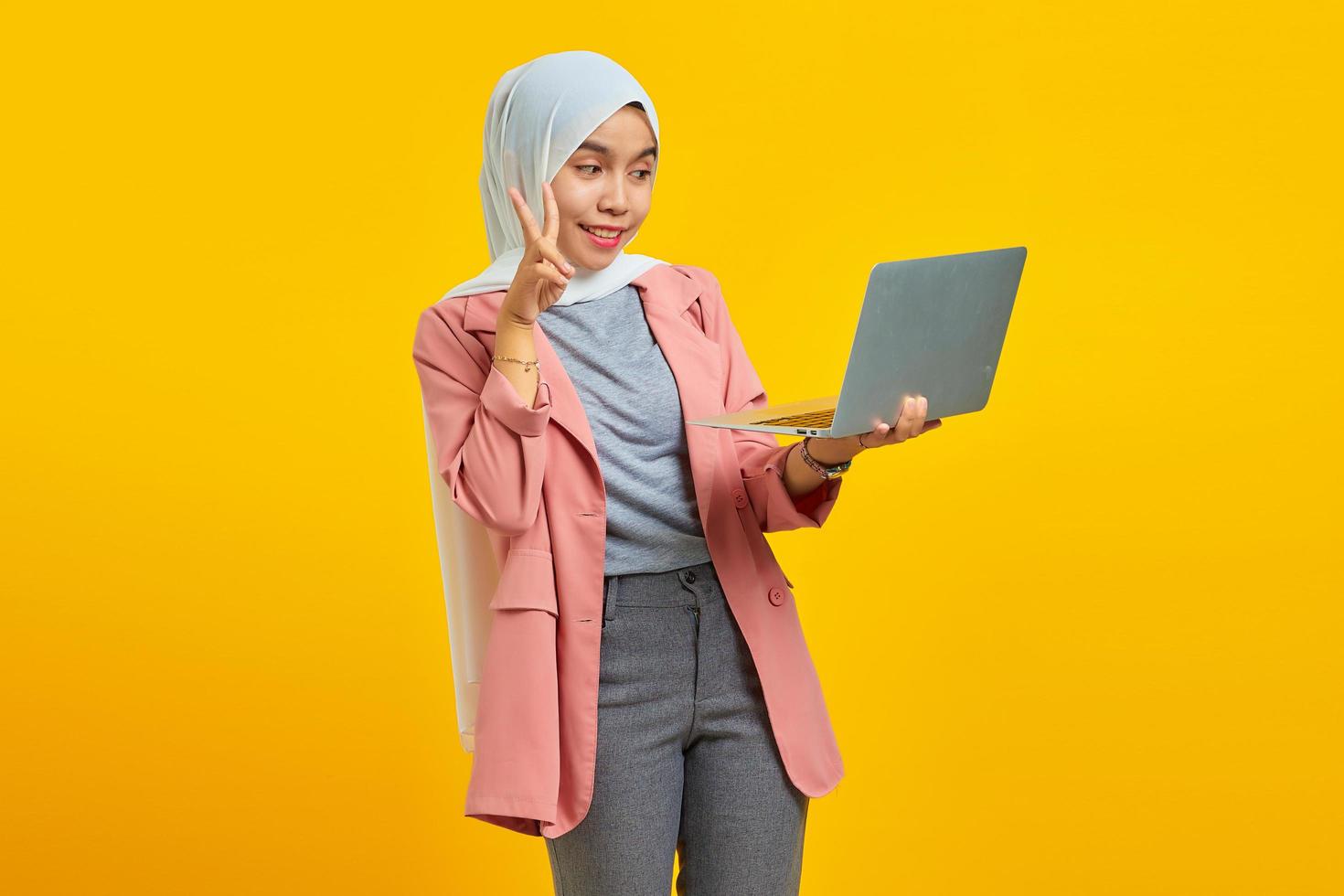 ritratto di bella giovane donna asiatica che tiene il computer portatile e fa segno di pace con le dita isolate su sfondo giallo foto