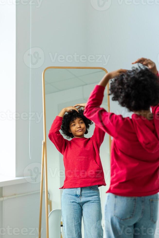 amore te stesso. bellissimo giovane sorridente africano americano donna danza toccante Riccio capelli godendo sua specchio riflessione. nero signora guardare a specchio guardare fiducioso e contento. se stesso amore concetto foto