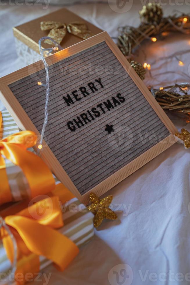 cartellone in feltro buon natale sul letto decorato con ghirlanda dorata, luci e scatole regalo foto