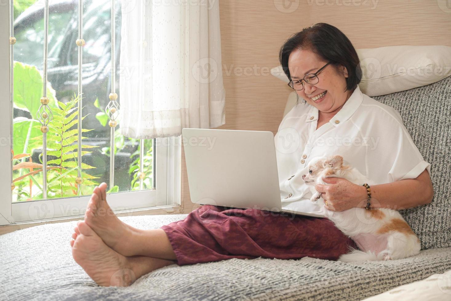 la donna asiatica anziana che si siede su un sofà sta usando un computer portatile. lei sorrise felice, il cane chihuahua si sedette sul lato. foto