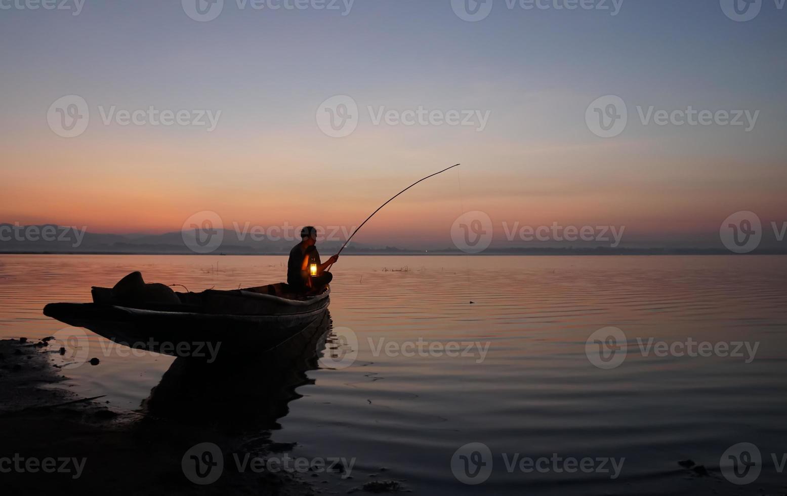 sul lato del lago, pescatore asiatico seduto sulla barca e usando la canna da pesca per catturare i pesci all'alba foto