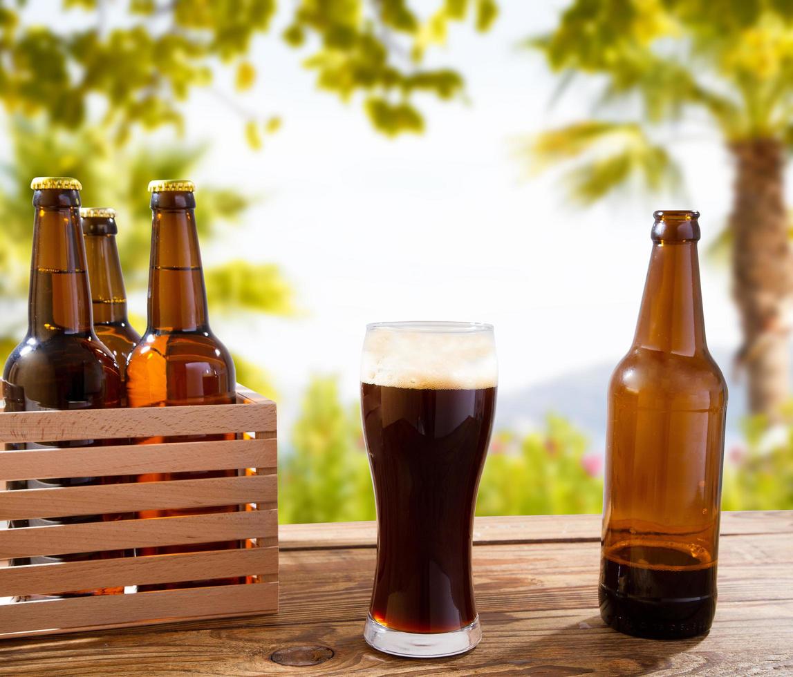 Bicchiere da birra scuro sul tavolo di legno con bottiglie,sfondo sfocato sulla spiaggia e sullo sfondo delle palme, concetto di cibo e bevande,spazio copia,messa a fuoco selettiva foto