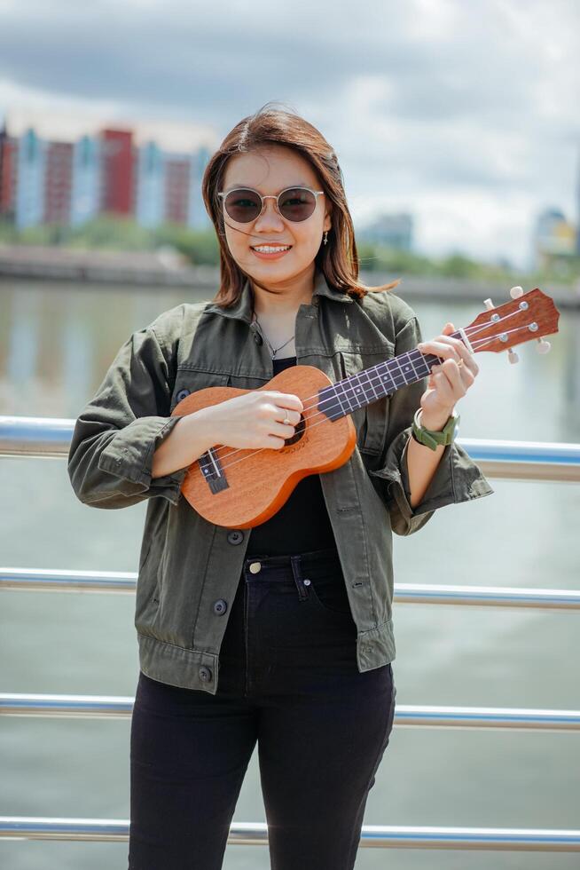 giocando ukulele di giovane bellissimo asiatico donna indossare giacca e nero jeans in posa all'aperto foto